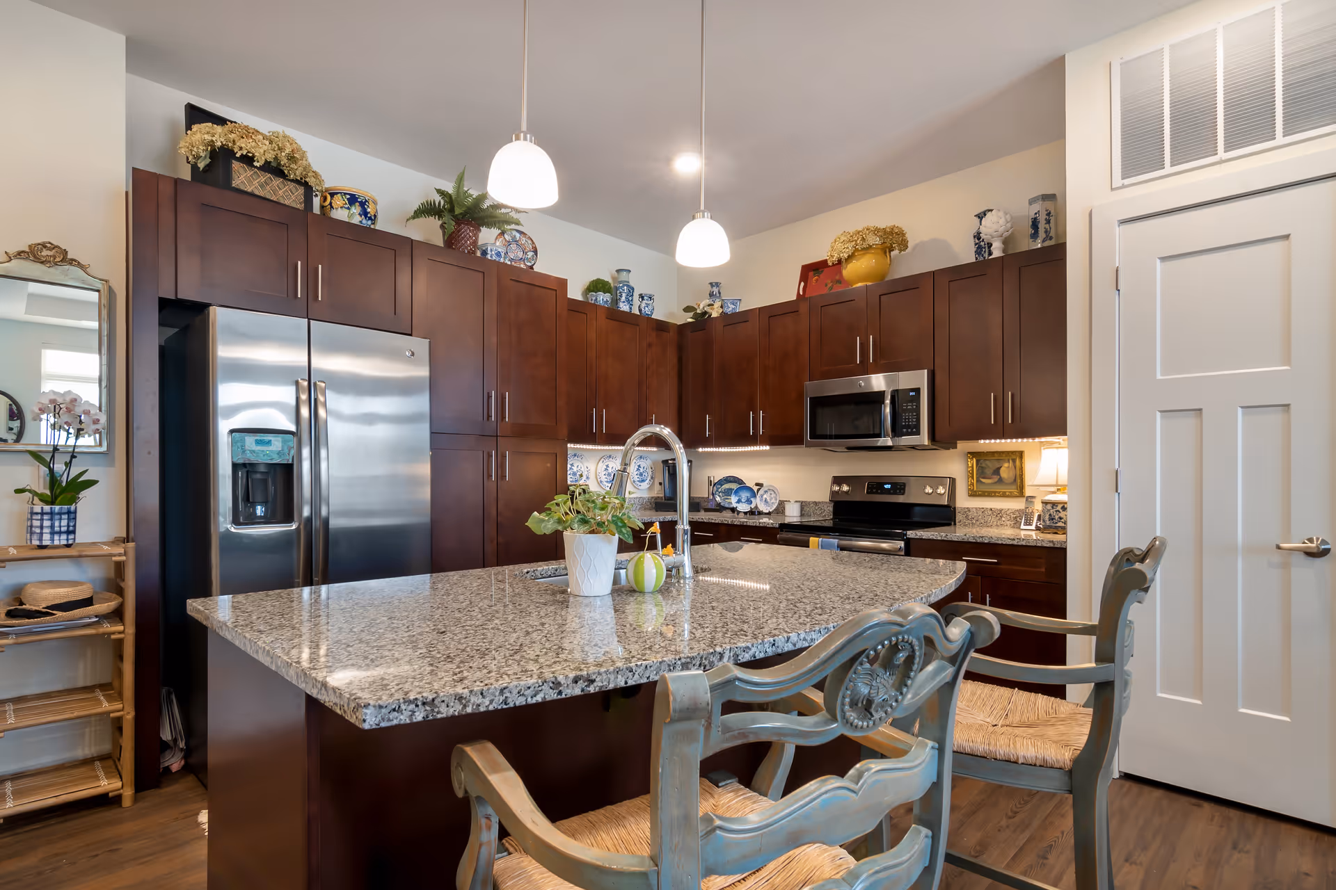 A modern kitchen with dark wooden cabinets, a large granite island countertop with a sink and two decorative chairs. Stainless steel refrigerator, microwave, and stove are visible. The kitchen is decorated with plants and ceramic plates on top of the cabinets.