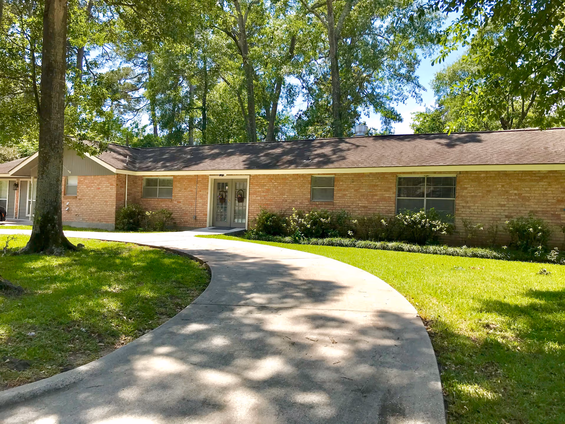Single-story brick building with a curved concrete walkway leading to double glass doors, surrounded by green grass, bushes, and tall trees under a clear blue sky.