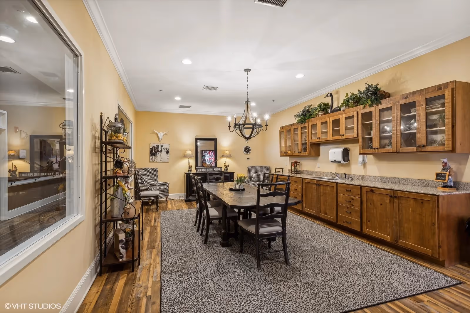 Communal dining room with a long wooden table and chairs, chandelier, built-in wooden cabinets and countertop, and decorative seating.