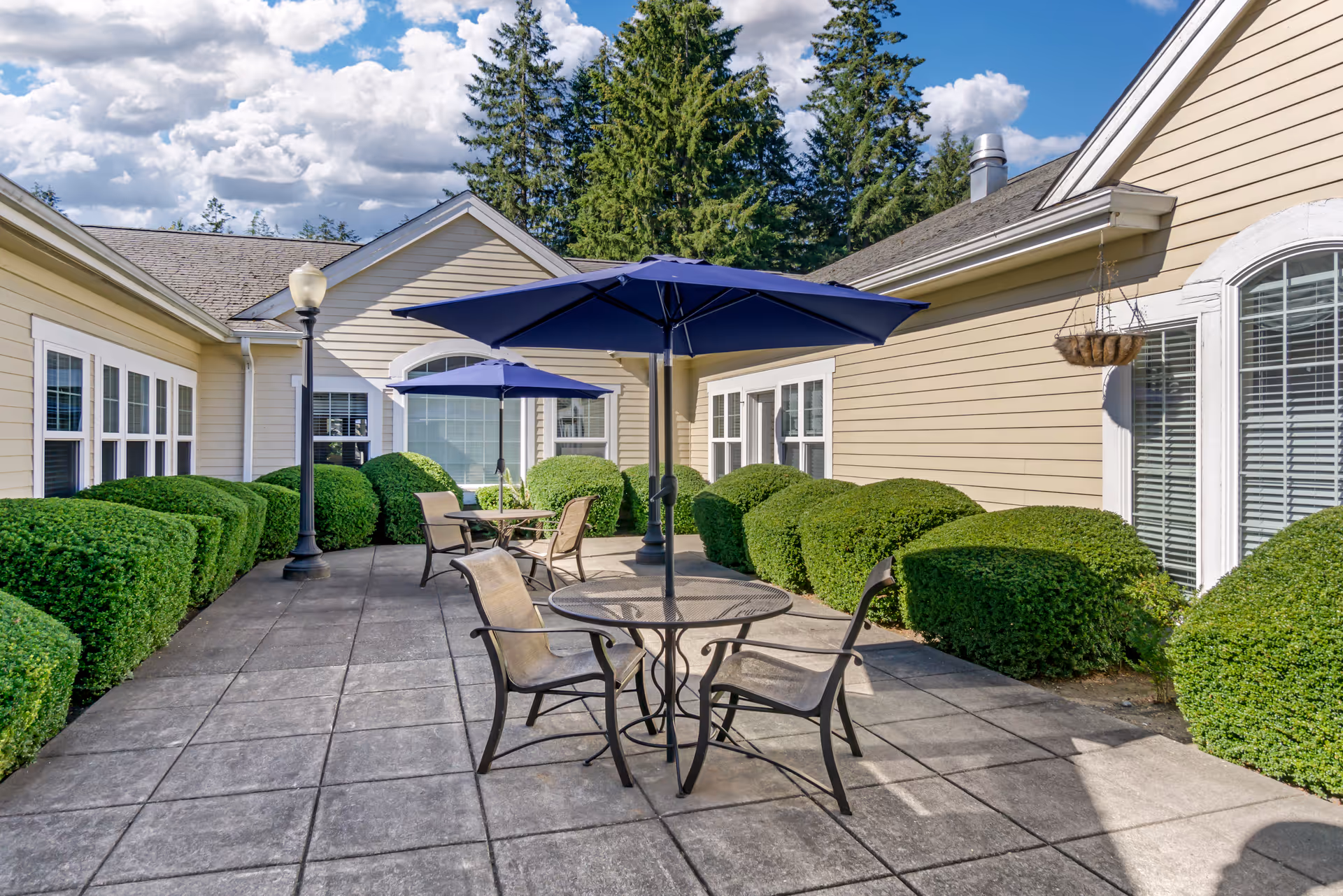 Outdoor patio area with two round metal tables, each with a large blue umbrella and four chairs. The patio is surrounded by neatly trimmed green bushes and beige siding buildings with white-framed windows. Tall evergreen trees and a partly cloudy sky are visible in the background.