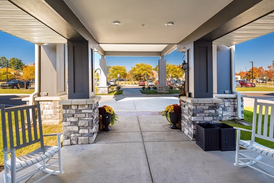 Covered entrance area of a senior living facility with stone pillars and two white rocking chairs on either side. There are potted plants with flowers near the pillars and a parking lot with cars and trees in the background under a clear blue sky.