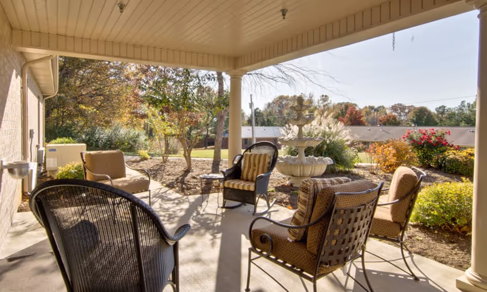 Covered outdoor patio area with several cushioned chairs arranged around a small table, overlooking a garden with a multi-tiered water fountain and various shrubs and trees under a clear sky.