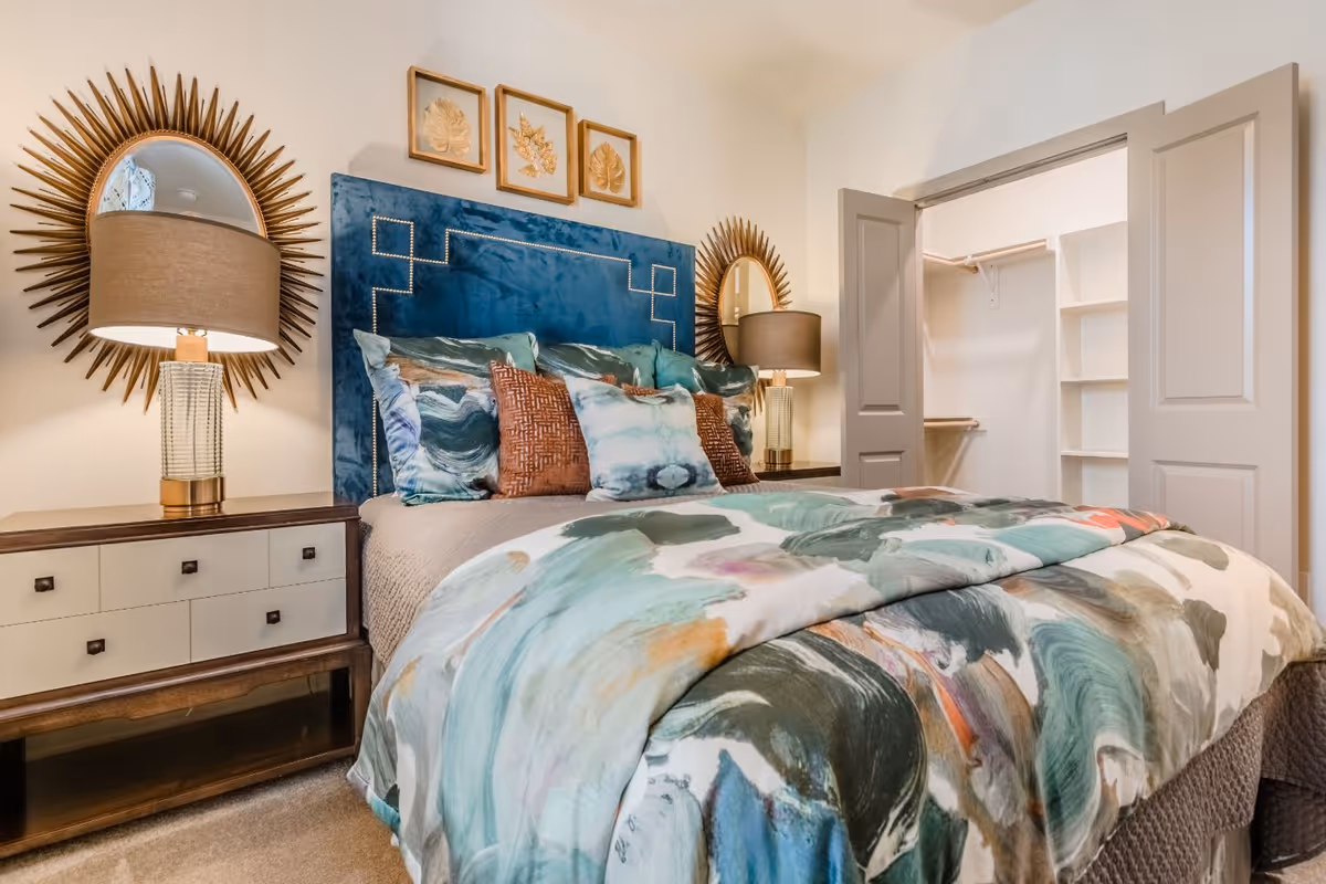 A cozy bedroom featuring a bed with a blue upholstered headboard and colorful bedding in shades of teal, brown, and white. On either side of the bed are matching nightstands with table lamps that have beige shades. Above the headboard are three framed leaf artworks. To the right, there is an open closet with shelves and hanging rods.