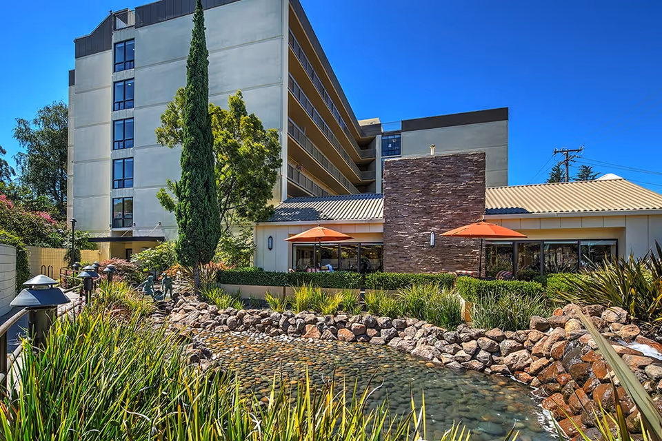 Exterior view of The Point At Rockridge Senior Living facility on a sunny day, showing a multi-story building with balconies, a landscaped garden with a pond and rocks in the foreground, tall trees, and two orange patio umbrellas near the building.