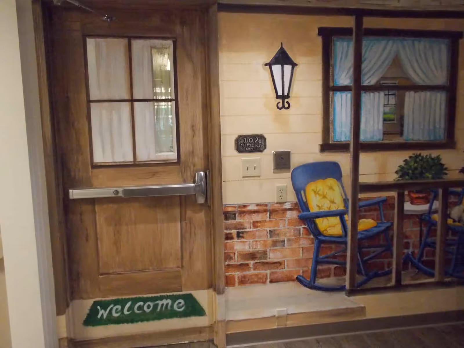 A painted wall mural depicting a wooden door with a window and a green welcome mat, a porch light, a sign reading '102 Memory Care', and a blue rocking chair with a yellow cushion next to a window with white curtains.