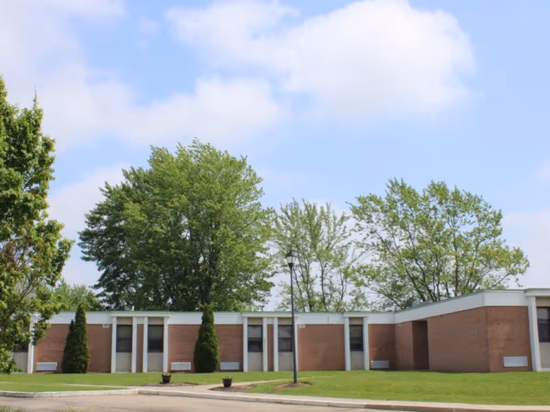 Single-story brick building with a grassy front lawn, trees, and a lamppost under a partly cloudy sky.
