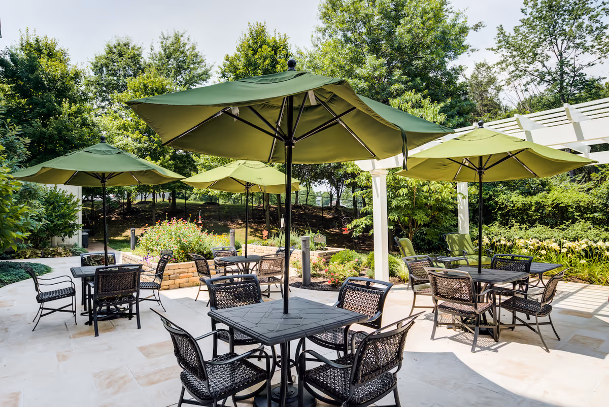 Outdoor patio area with several black metal tables and chairs, each table shaded by large green umbrellas. The patio is surrounded by lush greenery and trees, with a white pergola visible on the right side.