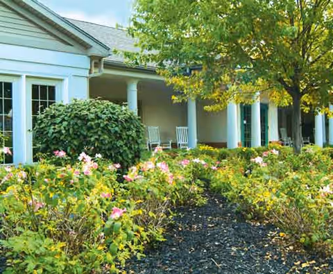 Front exterior of a single-story building with a columned porch, rocking chairs, and a flowering garden.