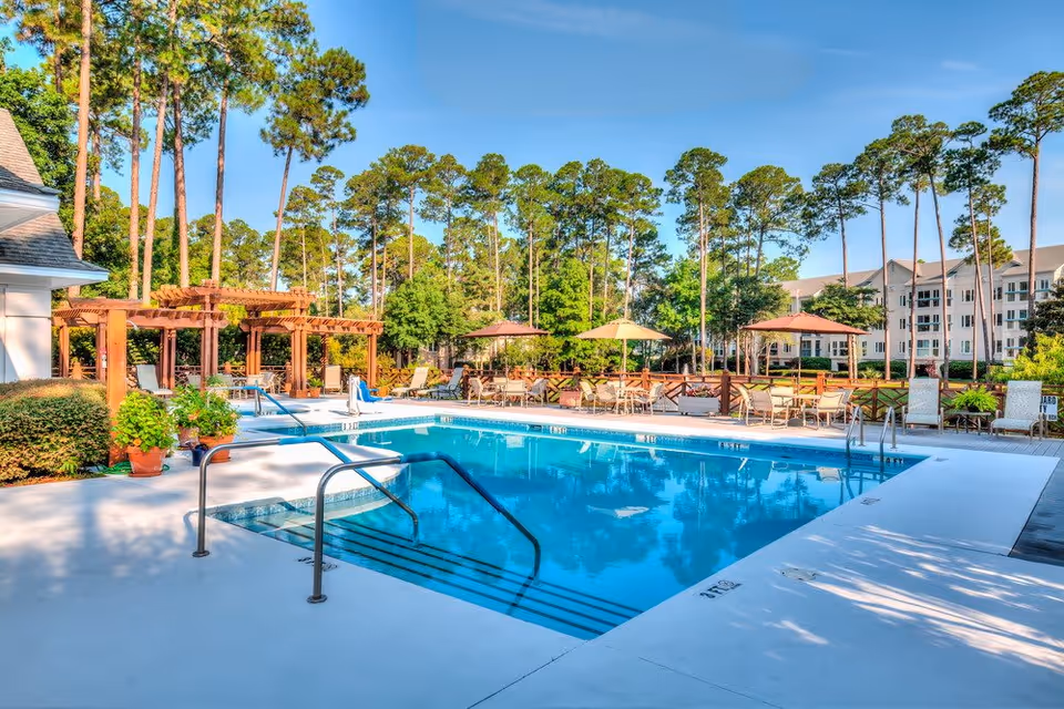 Outdoor swimming pool surrounded by lounge chairs, tables with umbrellas, and a wooden pergola. Tall pine trees and a multi-story residential building are visible in the background under a clear blue sky.