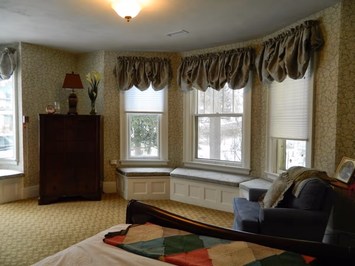 A cozy bedroom with a bed partially visible in the foreground, a dark wooden dresser with a lamp and vase on top, three large windows with decorative curtains and window seats beneath them, and a comfortable armchair with pillows and a throw blanket. The room has patterned wallpaper and carpet.