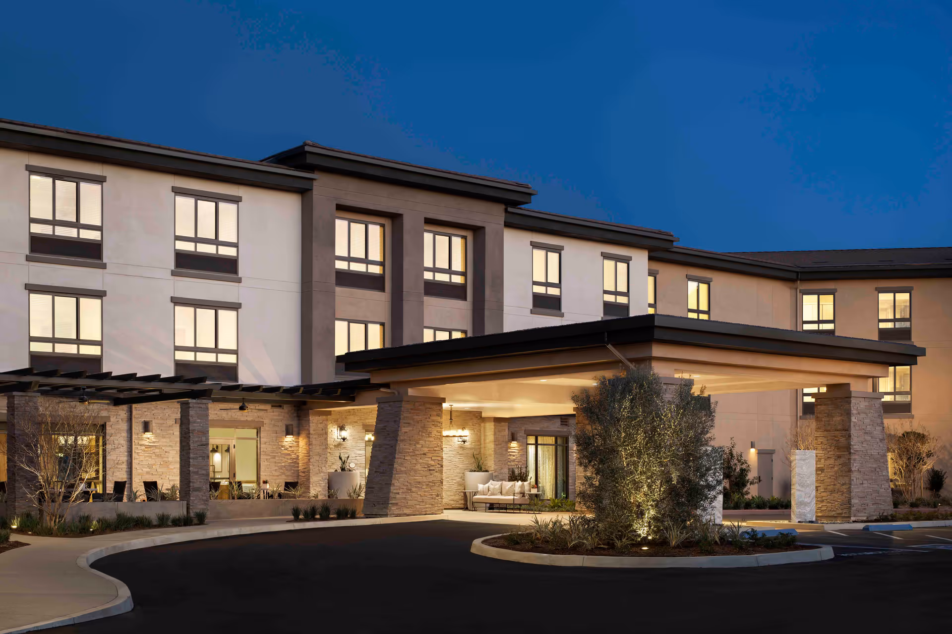 Exterior front entrance of a modern multi-story senior living building with a covered porte-cochere lit at dusk.