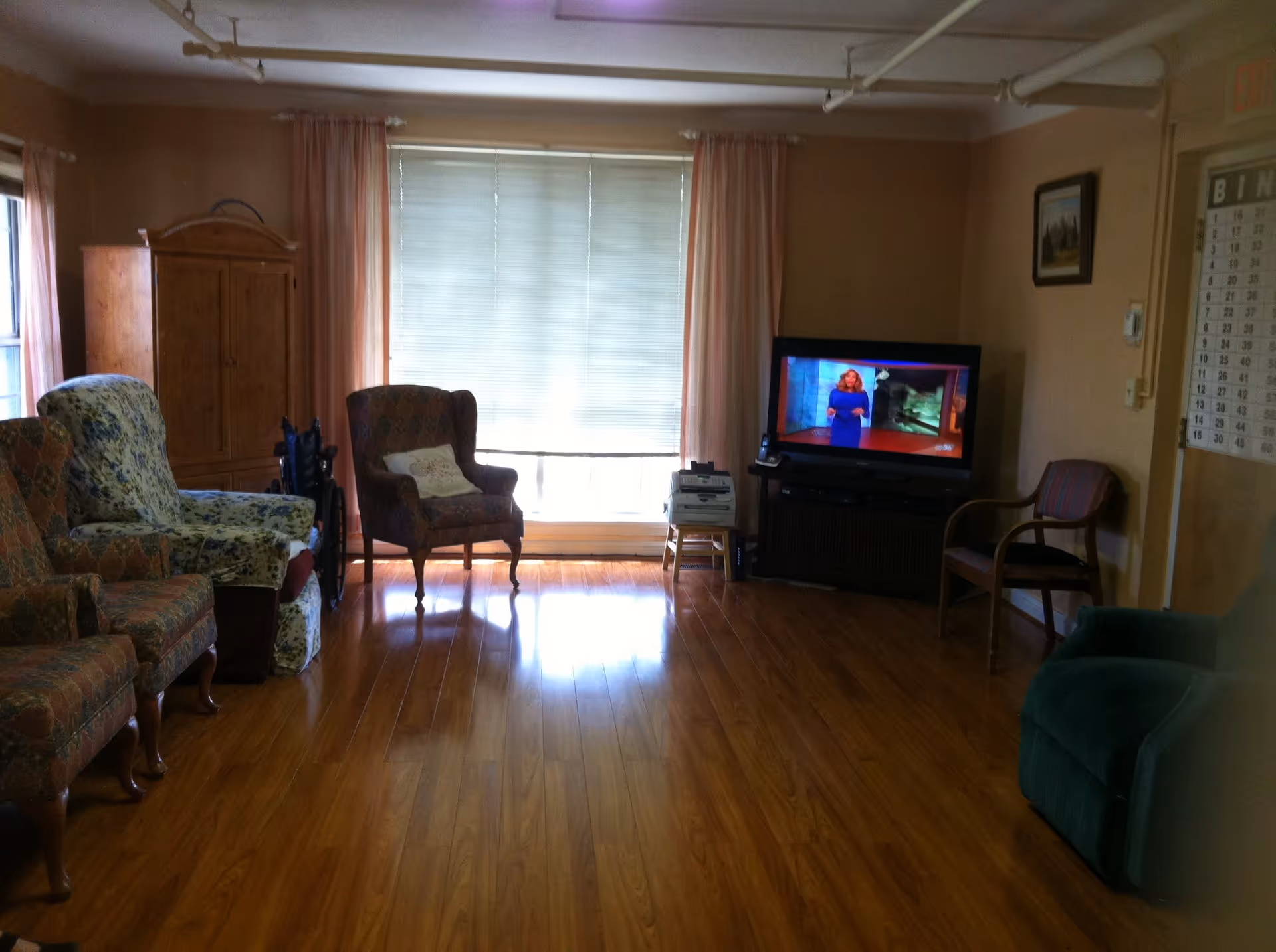 A living room with wooden flooring, several upholstered armchairs arranged along the walls, a television on a stand showing a woman in a blue dress, a wooden cabinet, and a large window with closed blinds and pink curtains.