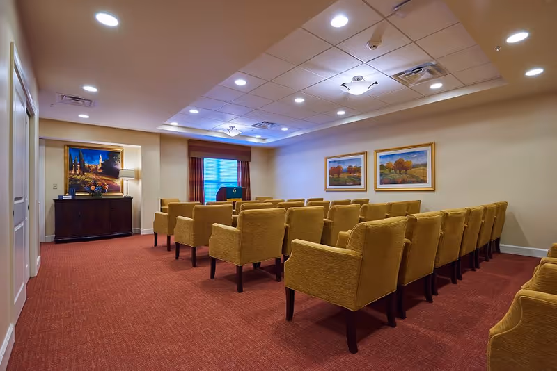A small conference or meeting room with rows of mustard yellow upholstered chairs facing a podium near a window with closed blinds. The room has red carpet flooring, beige walls, and a ceiling with recessed lighting and two framed landscape paintings on the wall.