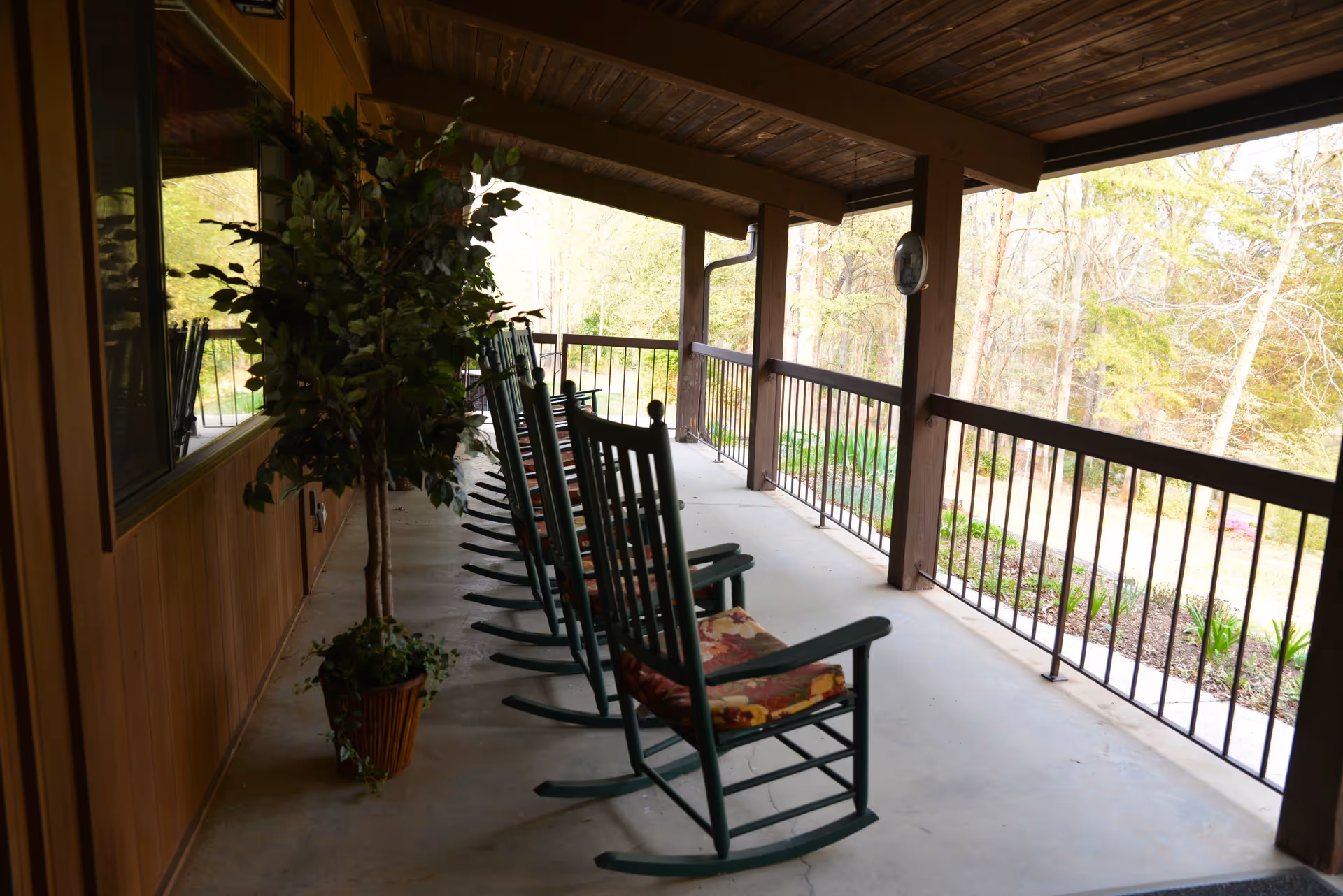 Covered outdoor porch with a row of green rocking chairs with floral cushions facing a wooded garden area. There is a potted plant near the wooden wall of the building.
