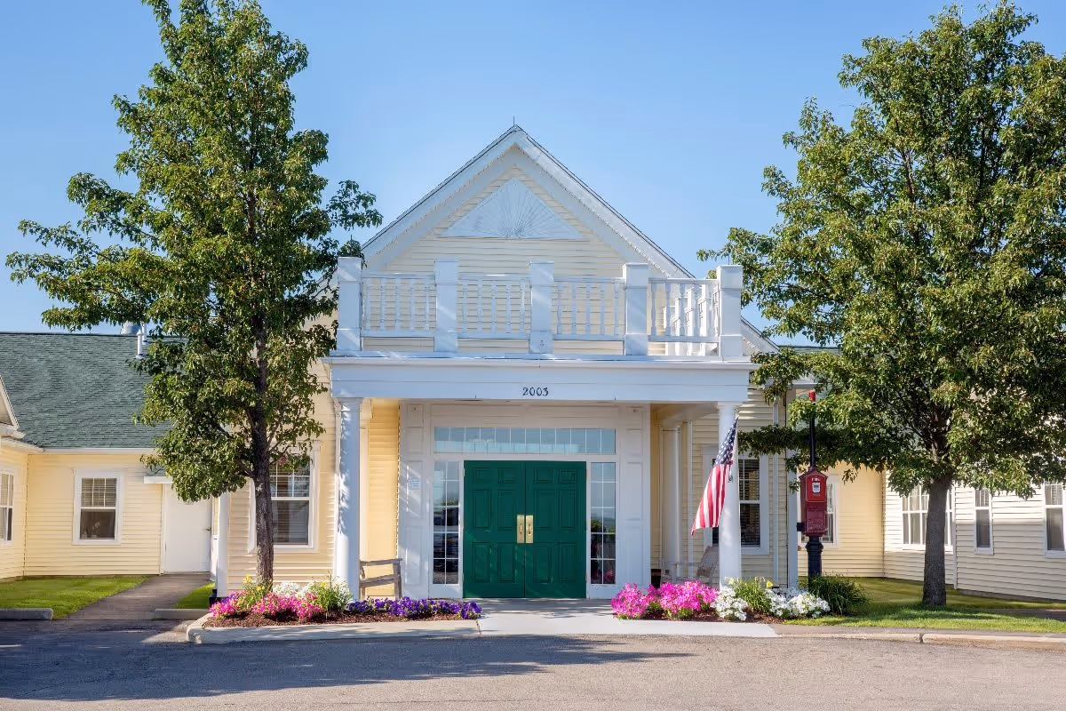 Front exterior view of a yellow building with a green double door entrance, white columns, and a small balcony above the entrance. There are two trees on either side of the entrance, flower beds with colorful flowers, an American flag, and a red mailbox or fire alarm box near the right tree. The building number 2003 is visible above the door.