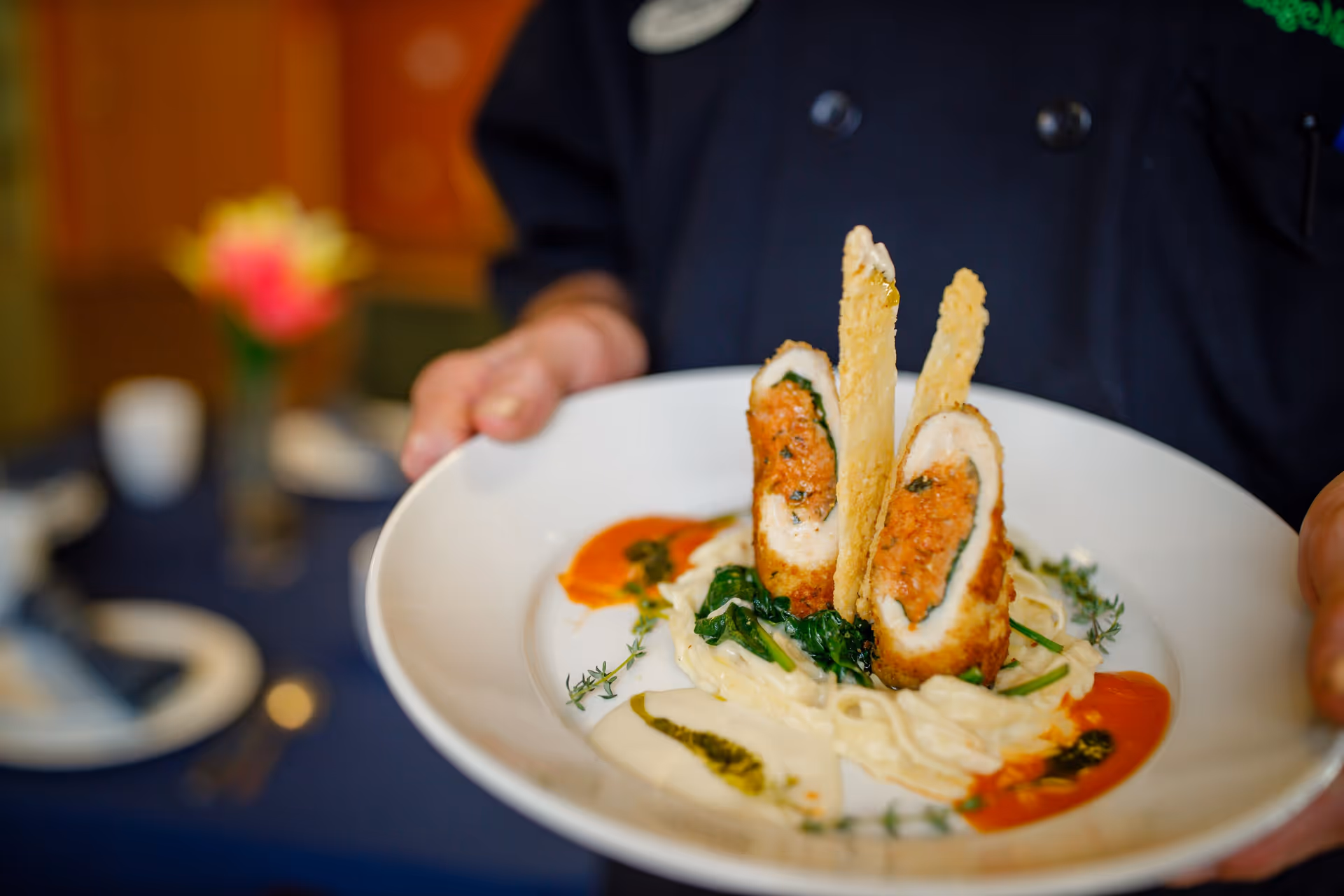 Person holding a white plate with a plated gourmet stuffed chicken roulade atop pasta with greens and sauces.