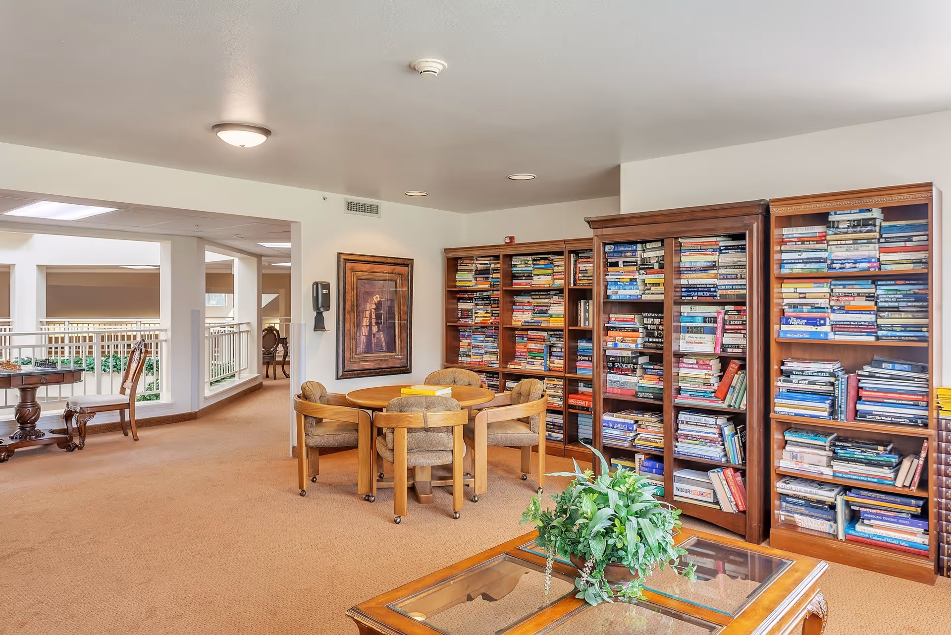 A cozy interior common area with carpeted floors featuring several wooden bookshelves filled with books, a round wooden table surrounded by four cushioned chairs, a glass coffee table with a green potted plant, and a framed artwork on the wall. The space is well-lit with ceiling lights and has an open view into an adjacent area with additional seating and railings.