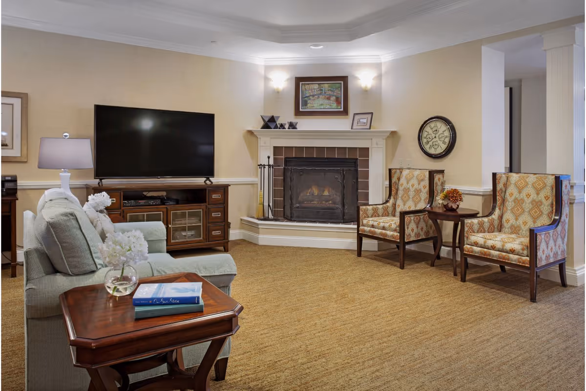 A cozy living room with a beige carpet and light-colored walls. There is a light green sofa with white flowers on a wooden side table in the foreground. A large flat-screen TV sits on a wooden cabinet against the left wall. In the center background is a fireplace with a fire burning, flanked by two wall sconces and decorated with a framed painting and small decorative items on the mantel. To the right, two patterned armchairs flank a small round wooden table with a flower arrangement. A large round clock hangs on the wall above the chairs.