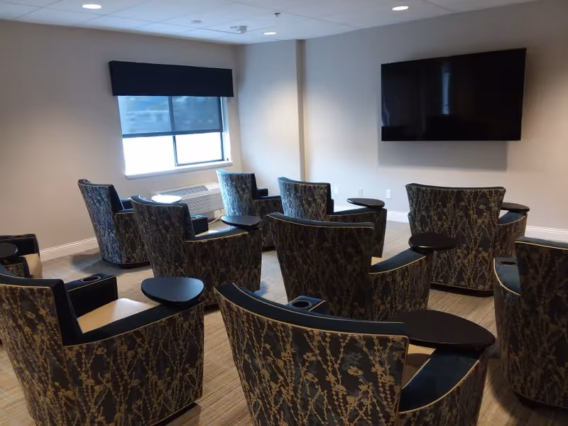 A small theater or media room with patterned armchairs arranged in rows facing a large flat-screen TV mounted on a beige wall. There is a window with a dark valance on the left side and a carpeted floor.