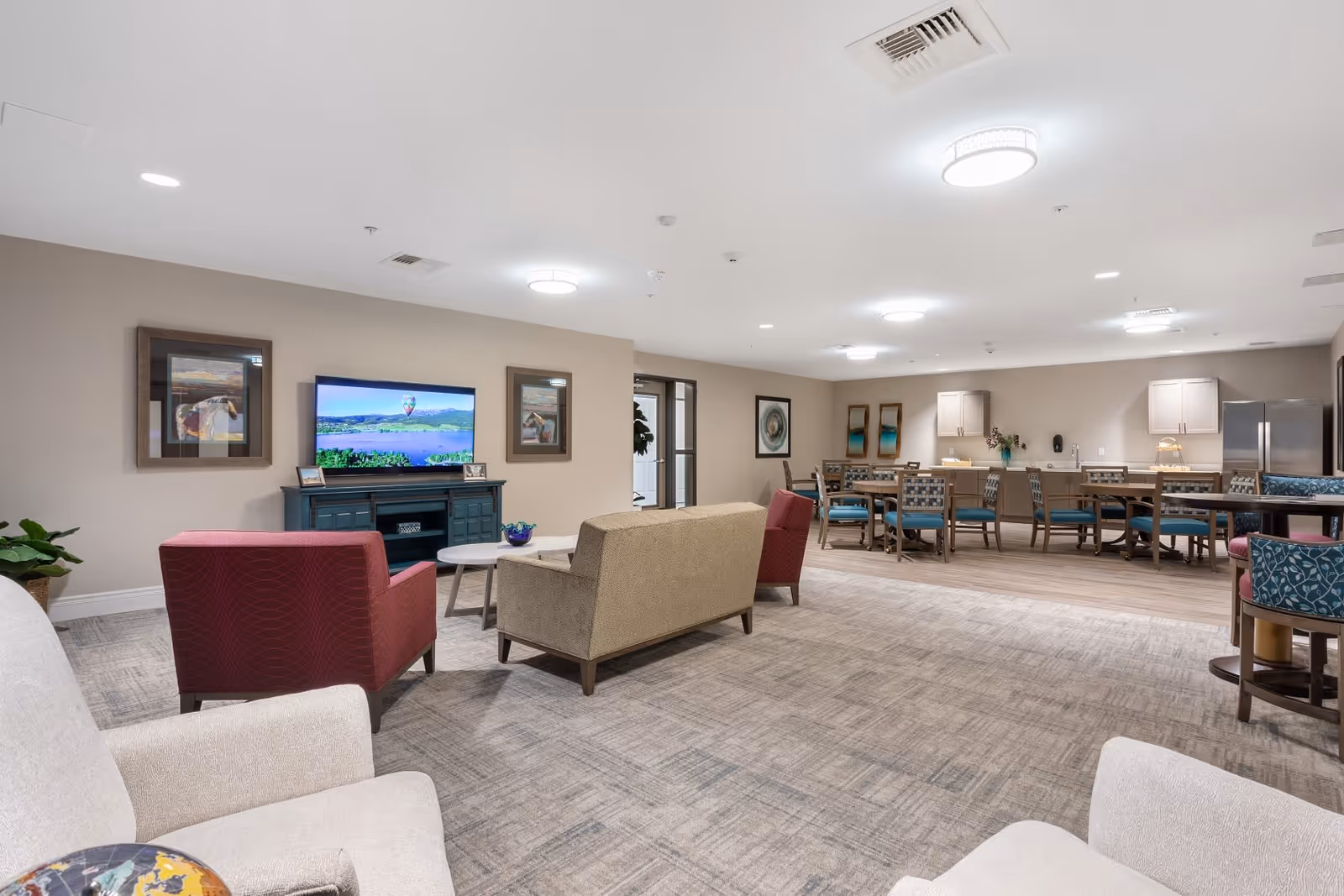 A spacious senior living common area featuring comfortable seating including armchairs and sofas arranged around a coffee table and a television mounted on a blue console. The room has neutral-colored walls adorned with framed artwork, and the far side includes a dining area with multiple tables and chairs, cabinetry, and a refrigerator. The space is well-lit with ceiling lights and has a carpeted floor transitioning to wood flooring in the dining area.