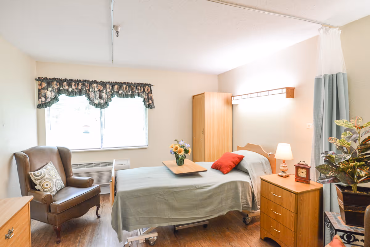 A well-lit bedroom in a senior living facility featuring a single bed with a green bedspread and a red pillow. There is a wooden bedside table with a lamp and a small clock, a wooden wardrobe, a brown armchair with a decorative pillow, and a window with floral curtains. A tray with a vase of flowers is placed on the bed, and a privacy curtain is partially visible on the right side.