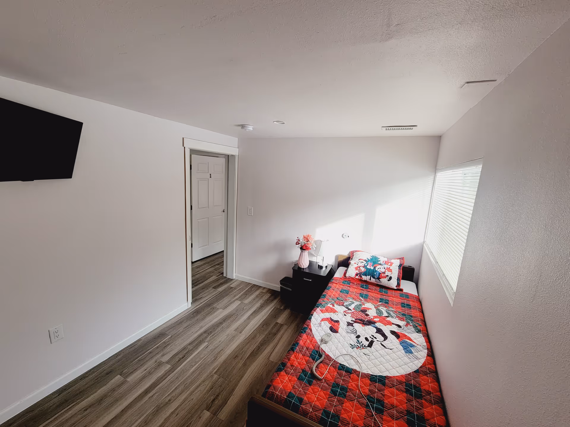 A small, bright bedroom with a single bed covered in a colorful quilt featuring cartoon characters. There is a window with blinds on the right wall letting in natural light. Next to the bed is a black nightstand with a pink vase of flowers and a small lamp. A flat-screen TV is mounted on the left wall, and a door is visible leading to another room.