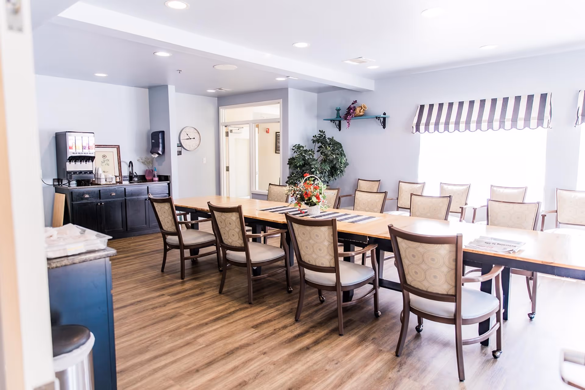 Bright communal dining room with long wooden tables, upholstered chairs, a floral centerpiece, and a beverage station.