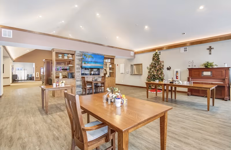 Spacious senior living common area with wooden dining tables and chairs, a TV and shelving unit, a decorated Christmas tree, and a piano.