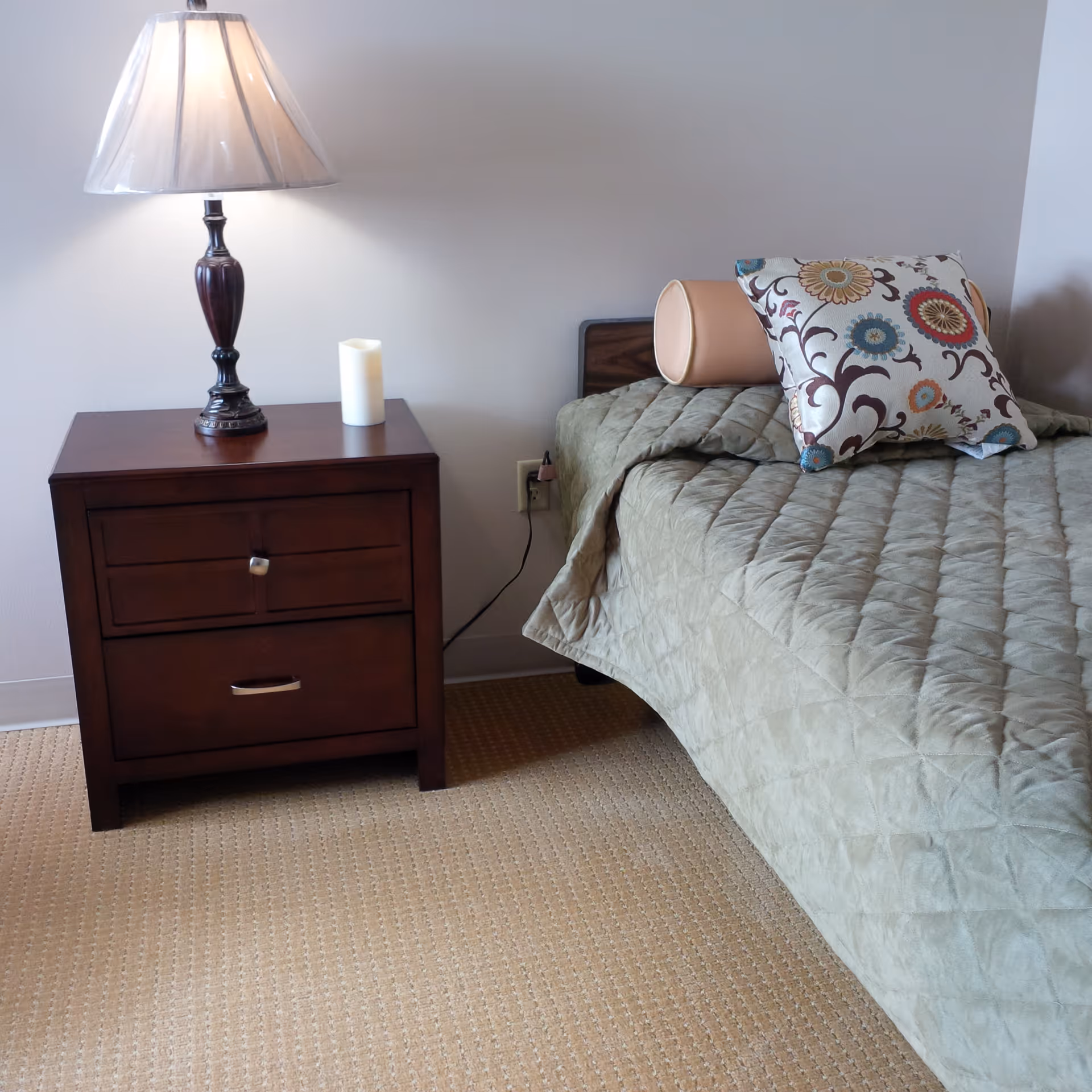 A bedroom corner featuring a wooden nightstand with two drawers, a decorative table lamp with a beige lampshade, and a small white candle. Next to the nightstand is a bed with a green quilted bedspread, a cylindrical bolster pillow, and a square pillow with a colorful floral pattern.