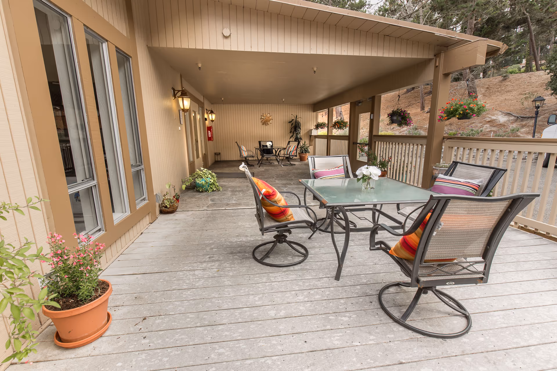 Covered outdoor patio area with a glass-top table and four chairs, each with colorful cushions. Several potted plants and hanging flower baskets decorate the space. The patio has wooden flooring and railings, with a beige building wall on one side and a hillside with trees in the background.