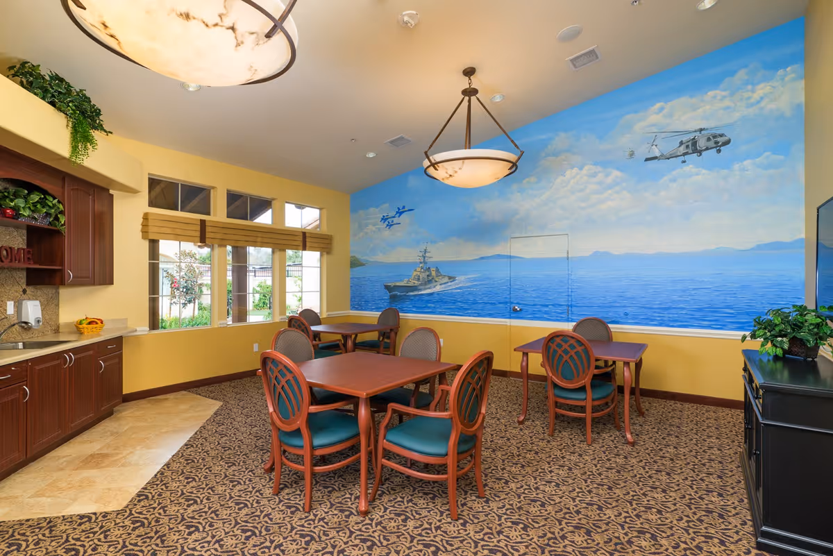 A bright and welcoming common area with several wooden tables and chairs arranged on a patterned carpet. The walls are painted yellow with a large mural depicting a naval scene including a ship, helicopter, and jets over the ocean. There are large windows with beige blinds letting in natural light, a countertop with cabinets and a sink on the left, and a black cabinet with a plant on the right. Two ceiling light fixtures provide additional lighting.