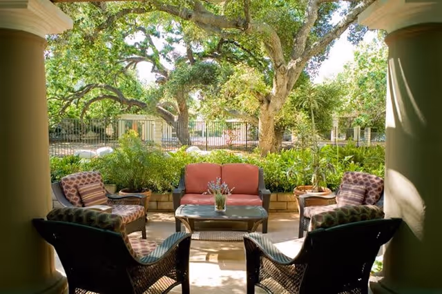 Outdoor patio seating area with cushioned wicker chairs and a loveseat arranged around a coffee table with a small plant, surrounded by lush greenery and large trees in the background.