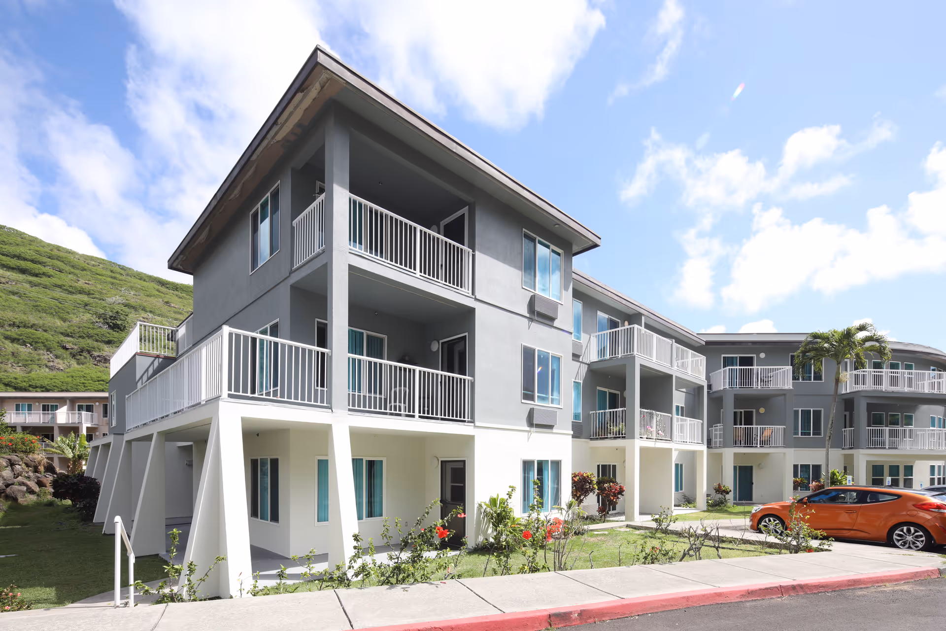 Exterior view of a modern three-story residential building with balconies, surrounded by greenery and a parked orange car in front. The sky is partly cloudy with a hill in the background.