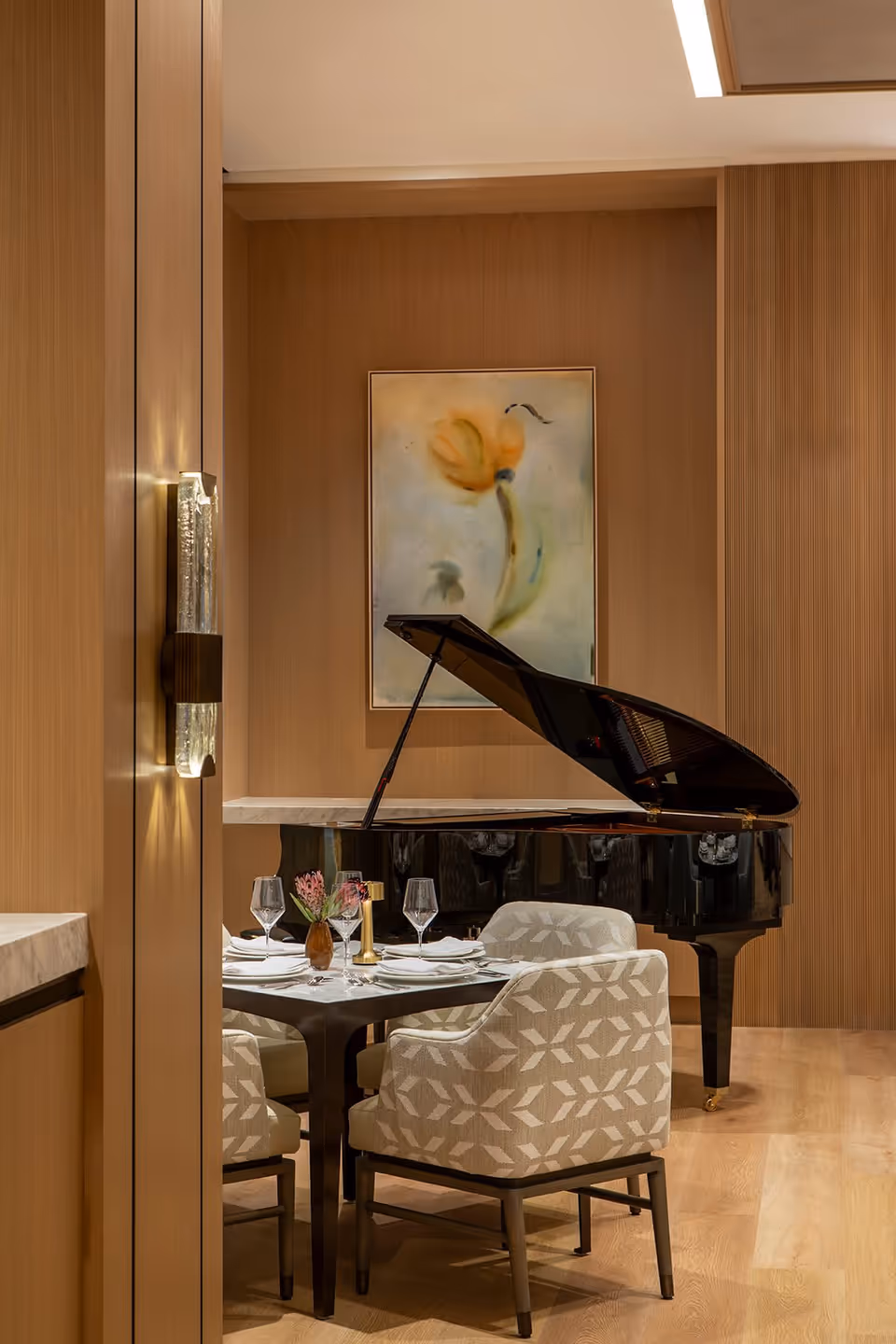 A cozy dining area with a small table set for four, featuring patterned upholstered chairs, wine glasses, and a small flower vase. Behind the table is a black grand piano with its lid open, and a framed abstract painting of a flower on the wooden-paneled wall.