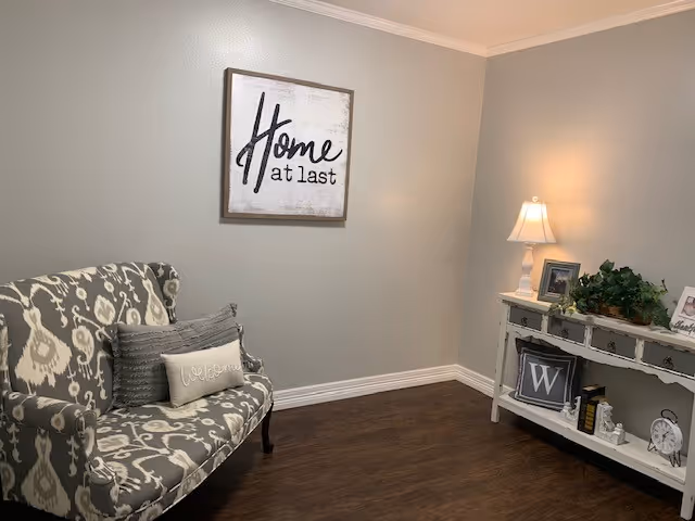 A cozy corner of a room with a patterned upholstered loveseat featuring two decorative pillows, one with the word 'welcome'. On the wall above the loveseat is a framed sign that reads 'Home at last'. To the right is a white console table with a table lamp, framed photos, a plant, books, and decorative items. The room has wooden flooring and light-colored walls.