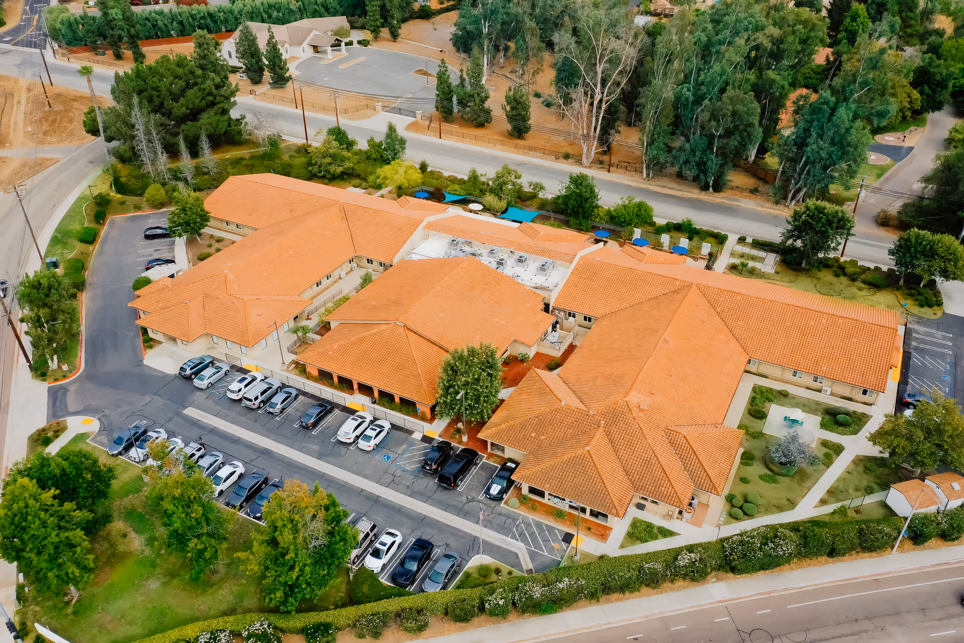 Aerial view of Vista Del Lago Memory Care facility showing a large building with orange tiled roofs surrounded by trees and greenery. There is a parking lot with cars parked in front of the building and roads surrounding the property.