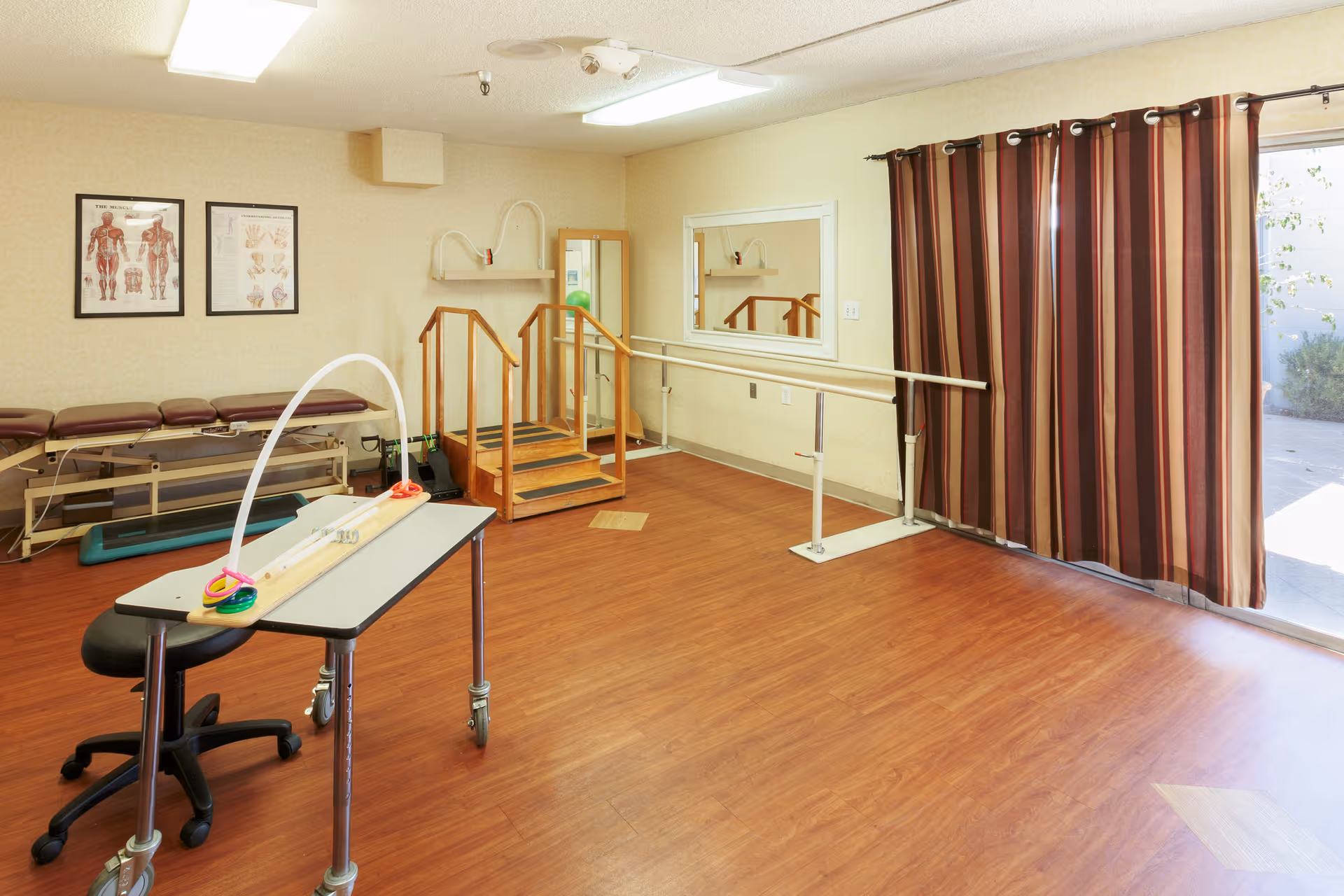 A therapy/rehab room with parallel bars, a small practice staircase, treatment tables, and a rolling therapy table near a curtained sliding door.