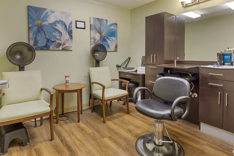Interior view of a salon area in a senior living facility featuring two green cushioned chairs with wooden arms, a round wooden table between them, two hair dryer chairs with hooded dryers, a black salon chair, and dark brown cabinetry with a large mirror above the counter. The walls are light green and decorated with two large blue floral paintings.