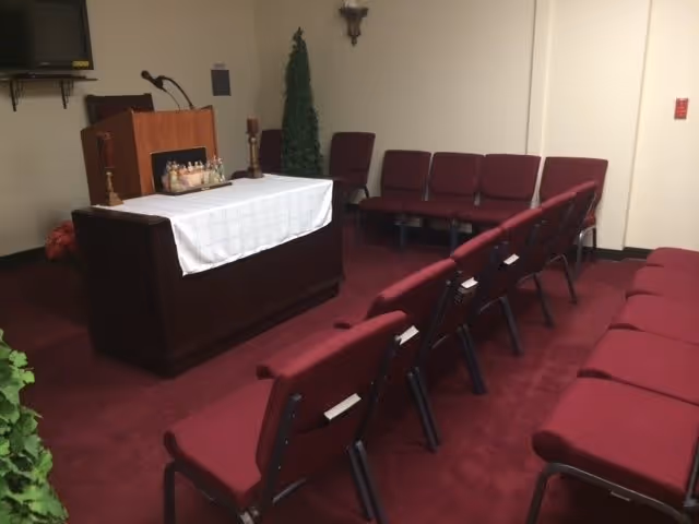 Small chapel or meeting room with rows of maroon chairs facing a lectern draped in a white cloth and a microphone.