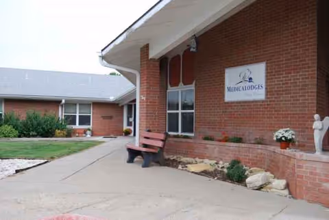 Front exterior of a brick senior living facility with bench, sign, and plants.