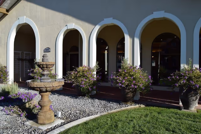 Outdoor courtyard area with a stone water fountain surrounded by purple flowering plants and greenery, in front of a building with arched doorways and windows.