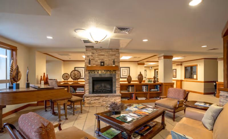 A cozy living room area in a senior living facility featuring a stone fireplace in the center, surrounded by wooden bookshelves with decorative vases and framed artwork. The room has comfortable seating including armchairs and a sofa, a wooden coffee table with magazines, and a piano near a window letting in natural light.