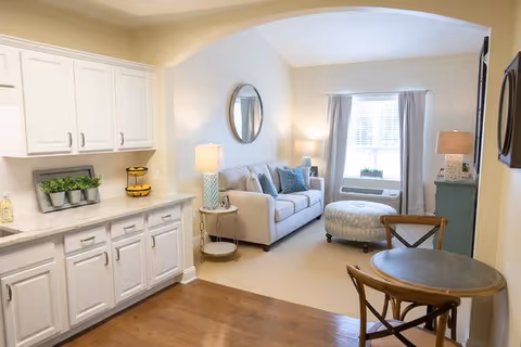 A bright and cozy living area in a senior living facility featuring a small round wooden dining table with two chairs, a beige sofa with blue and gray cushions, a round ottoman, a side table with a lamp, a round mirror on the wall, a window with curtains, and white kitchen cabinets with a countertop decorated with small potted plants and a tiered fruit basket.