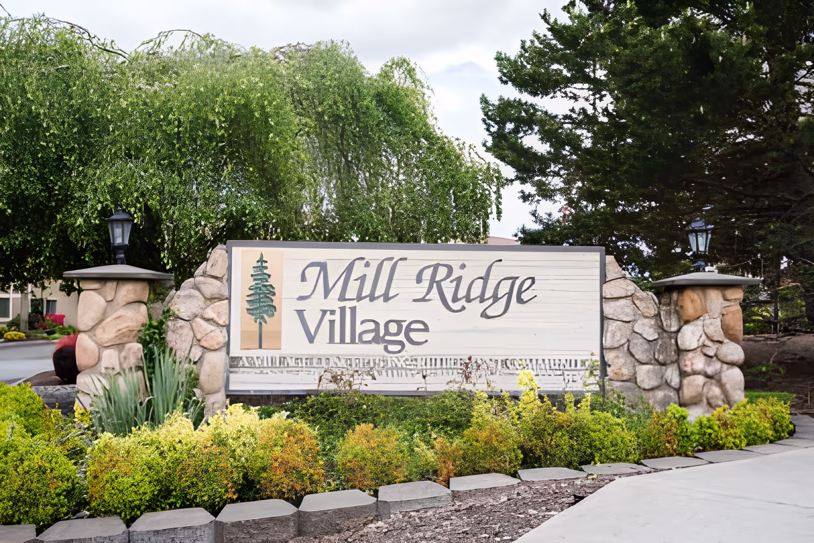 Stone entrance sign for Mill Ridge Village surrounded by green bushes and trees, with two stone pillars topped with lanterns on either side of the sign.