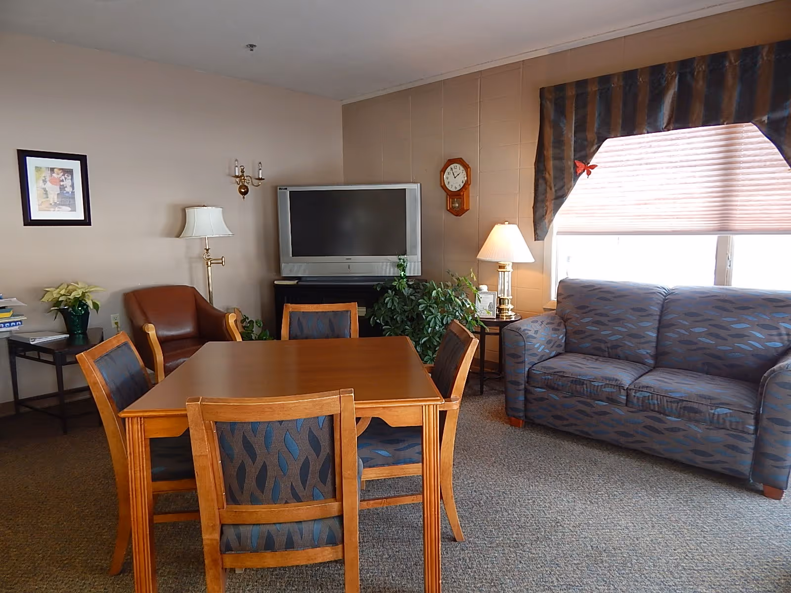 A cozy living room area with a wooden table and four chairs in the center. To the right, there is a patterned blue couch in front of a window with blinds and a striped valance. A floor lamp and a side table with a lamp and a plant are next to the couch. On the left side, there is a brown leather armchair, a small table with books and a plant, and a wall-mounted TV above a cabinet. A wall clock and framed picture are also visible on the walls.