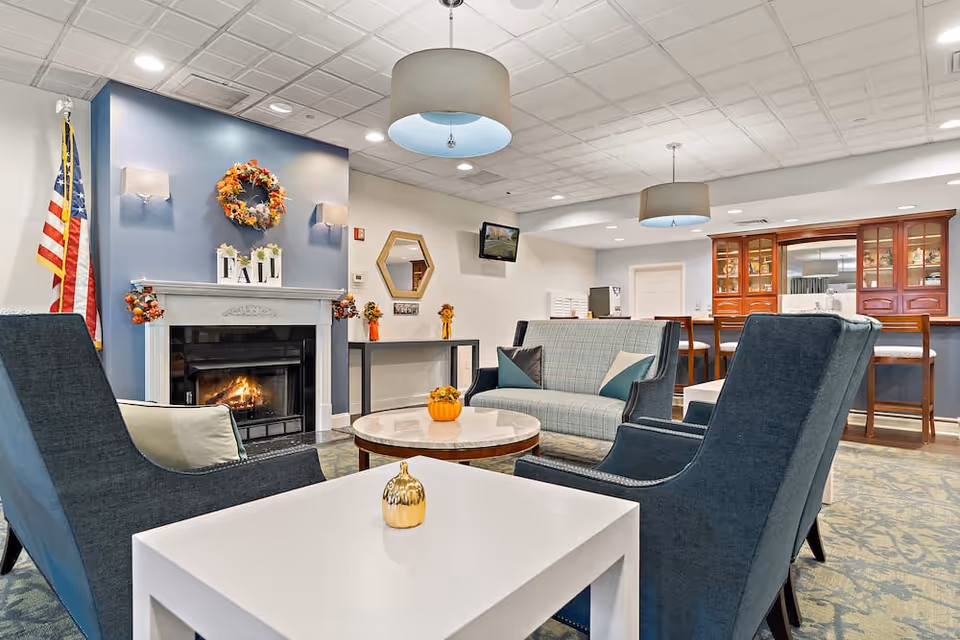 A cozy living room area in a senior living facility featuring a lit fireplace with fall decorations, including a wreath and small pumpkins. The room has comfortable seating with two blue armchairs, a plaid sofa, and a round coffee table. There are two pendant lights hanging from the ceiling, an American flag in the corner, a wall-mounted TV, and a wooden cabinet with glass doors in the background.