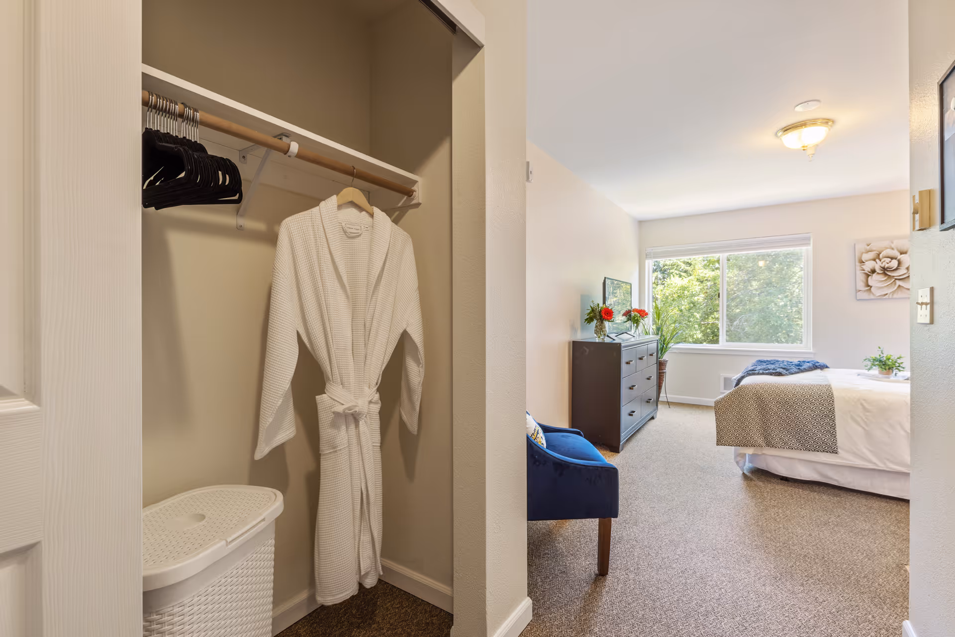 View of a bedroom from the entrance showing a closet with a white robe hanging and several black hangers. The bedroom has a large window with greenery outside, a bed with white and gray bedding, a dark dresser with flowers on top, a blue upholstered chair, and a beige carpeted floor.