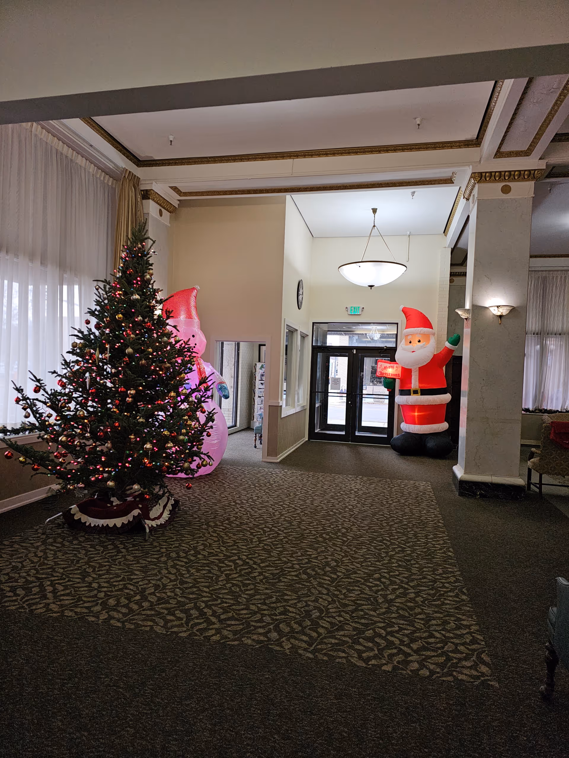 Interior view of a senior living facility lobby decorated for Christmas with a Christmas tree adorned with lights and ornaments on the left, and two large inflatable holiday figures, a snowman and Santa Claus, near the entrance doors. The space has patterned carpet, large windows with sheer curtains, and a hanging light fixture.
