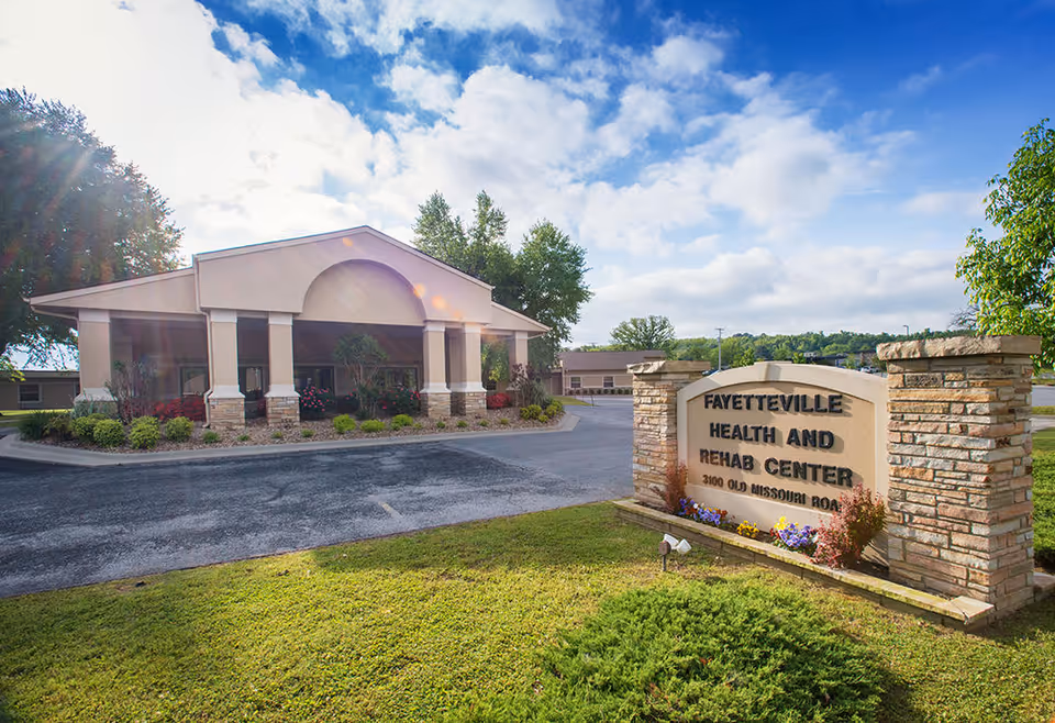 Exterior view of Fayetteville Health and Rehabilitation Center building with a covered entrance and a stone sign in front displaying the facility's name and address. The scene includes a paved driveway, green lawn, trees, and a partly cloudy sky.