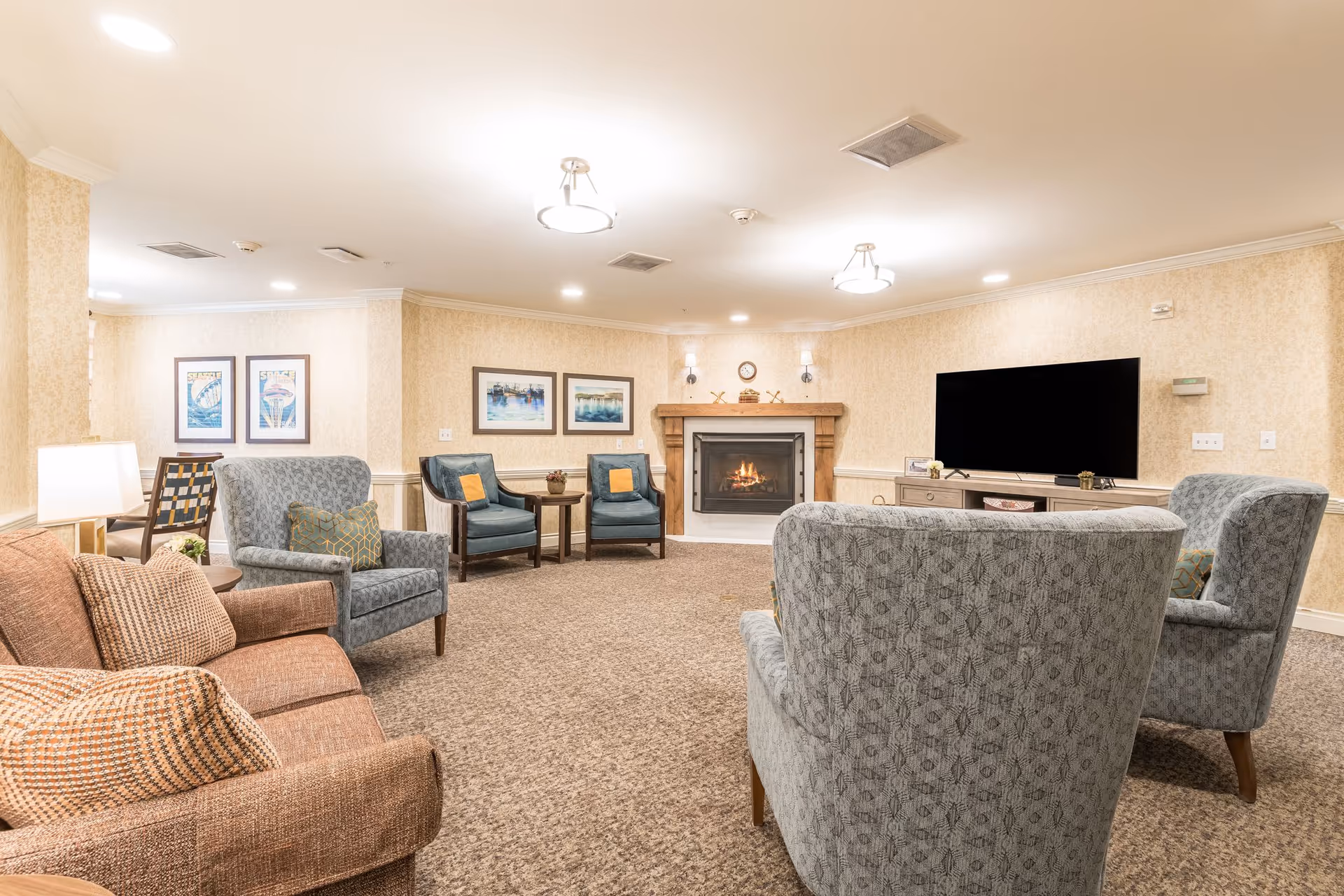 Cozy senior living common room with armchairs and sofas arranged around a fireplace and large wall-mounted TV.